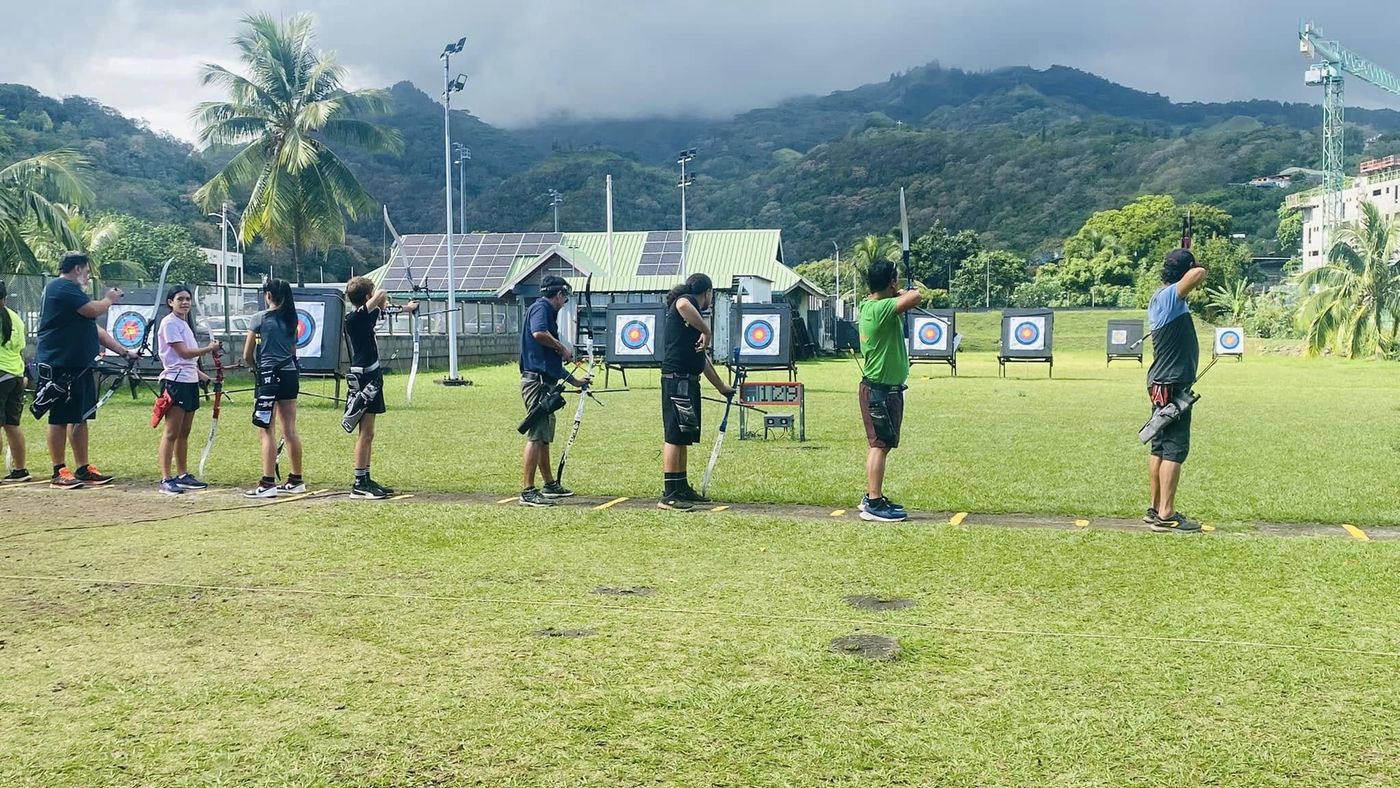 Archers du club de tous âges alignés sur la ligne de tir du stade Pater, face aux cibles, avec les cocotiers et les montagnes de Pirae en arrière-plan.